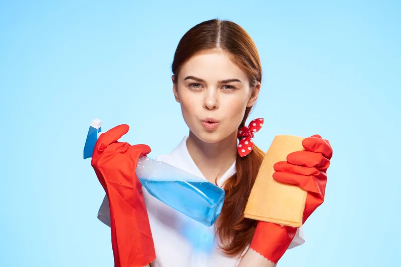 Весёлая уборщица с чистящими средствами на фоне голубого фона, оказывающая услуги по уходу за домом