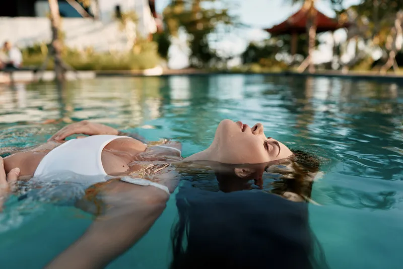 Relaxed poolside scene with a woman floating on water in a white bikini, eyes closed, calm expression, warm sunlight, tropical backdrop, a tranquil atmosphere of summer leisure