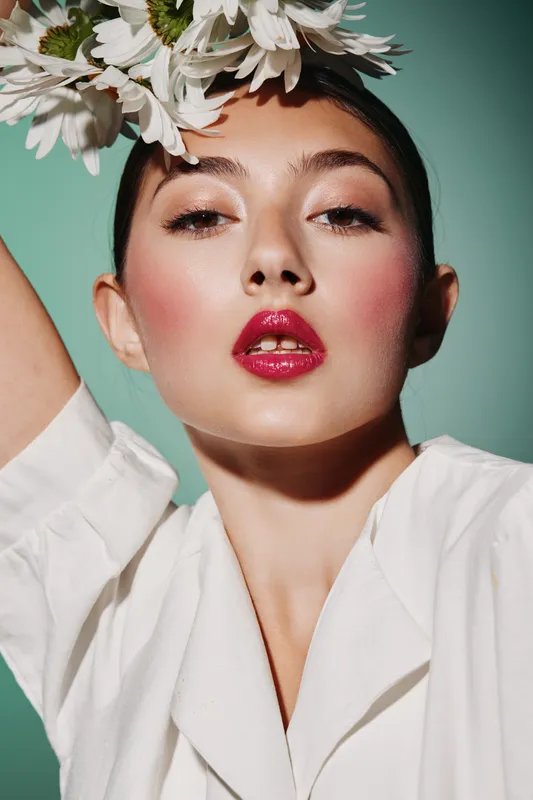 Close-up of a young woman with dewy skin and bold pink lips posing with white flowers on her head against a blue background. Creative beauty and fashion concept.