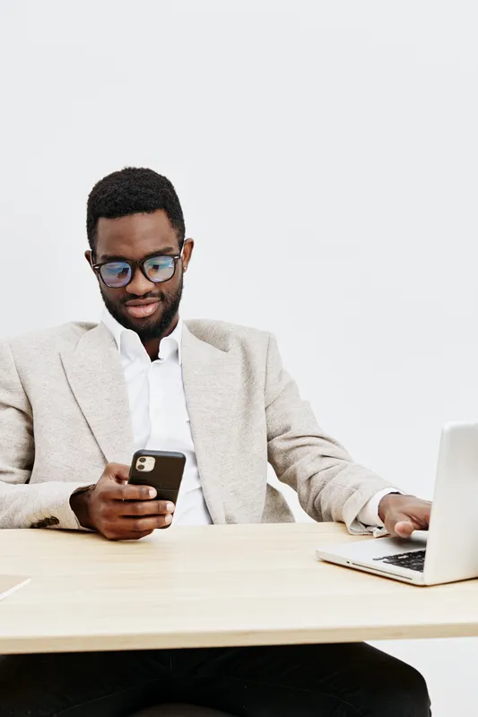 Professional young African American man working on a smartphone and laptop in a bright minimalistic office space. Business, technology, focus, productivity, communication, modern lifestyle.