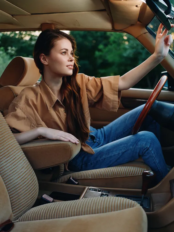 Young woman in stylish beige shirt and blue jeans sitting in vintage car, enjoying a moment of relaxation on a sunny day.