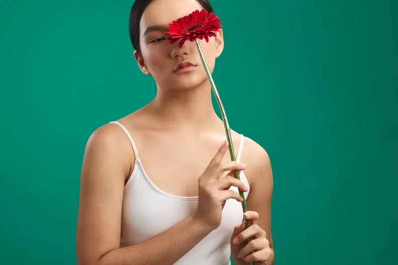 asian woman holds a red flower against a teal backdrop in a studio portrait, soft lighting highlights calm expression, minimalist attire, graceful pose.