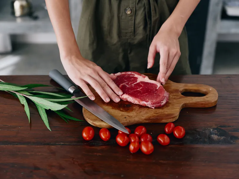 Женщина с ножом в руке нарезает свежее мясо стейка для жарки на кухне