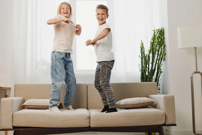 Happy kids jumping on sofa in bright living room, smiling and playing together. Concept of childhood fun, energetic siblings, and home leisure activity.