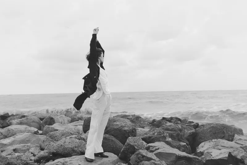 Woman on rocks by the sea raises arms in triumph, monochrome seascape with dramatic mood, conveying freedom, resilience and bold movement against the horizon