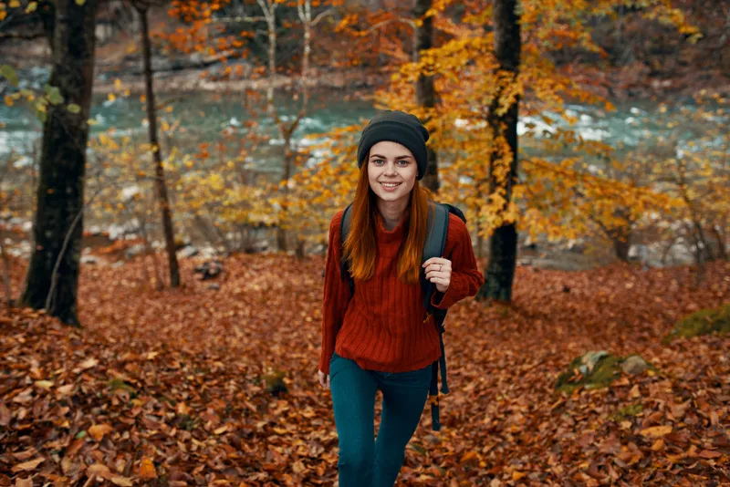 Путешественник в красном свитере и шапке идет по осеннему лесу возле горной реки, вид сверху