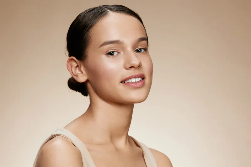 Young woman with natural makeup smiling, showcasing healthy skin tone against a warm, neutral background, ideal for beauty and skincare themes