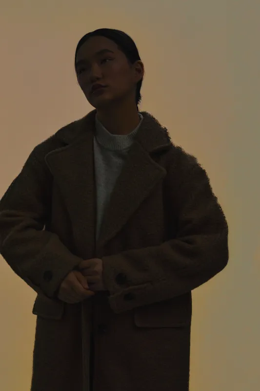 asian woman in a warm coat stands in a soft shadowed setting, conveying introspection and calm as moody lighting creates an intimate portrait.
