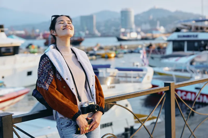 Woman in a patterned cardigan and scarf stands on a marina railing, harbor boats and water behind, basking in bright sunlight with a relaxed fashion vibe and joyful mood