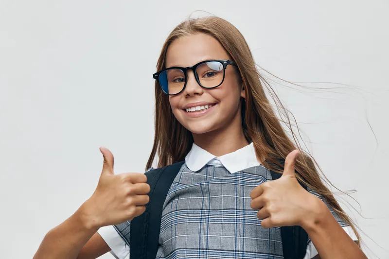 Girl with glasses smiling and showing thumbs up gesture, wearing checkered dress and white collar against light background in studio portrait, positive and confident expression.