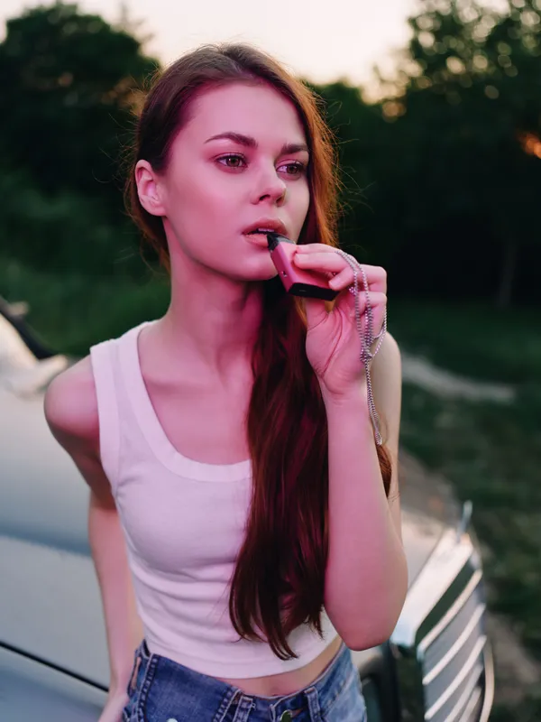 Young woman with long hair, wearing a white tank top and denim shorts, holding a whistle, exuding confidence against a vibrant sunset backdrop.