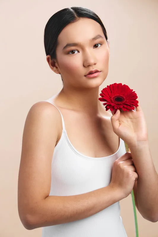 Asian woman stands gracefully in a soft studio setting, holding a red flower close to her face. She gazes softly at the camera, exuding calm confidence and subtle elegance.