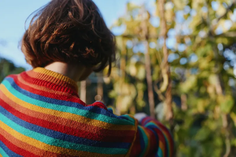 Woman in striped sweater seen from back view in a garden outdoors, short hair, casual outfit, reaching among plants, relaxed lifestyle scene for fashion and travel imagery.