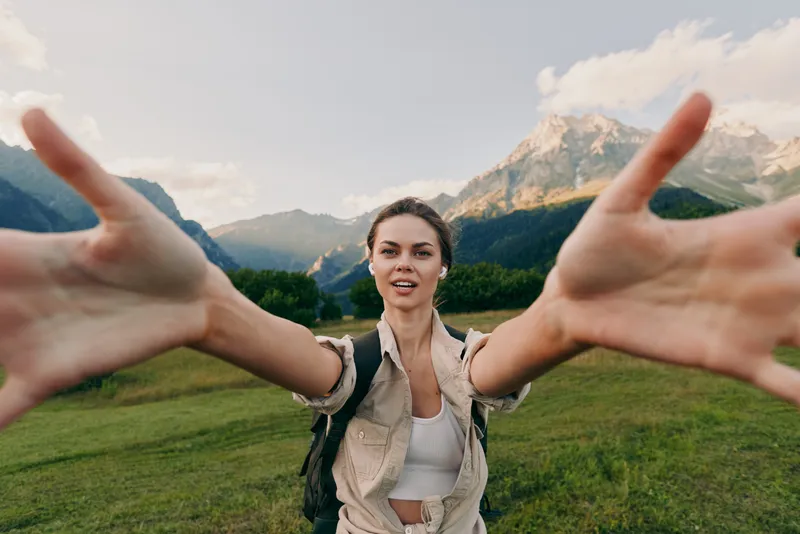 A young woman in casual clothes stretches her arms forward smiling in a green mountain meadow under a blue sky. Concept for lifestyle, nature, and freedom.