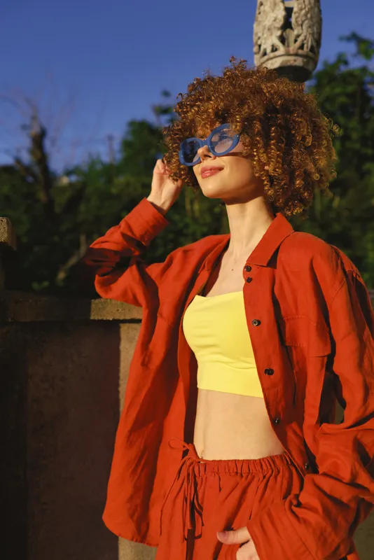 Young woman with curly red hair stands outdoors in sunny weather, wearing a bright yellow crop top and orange jacket, stylish sunglasses, casual fashion lifestyle pose amid warm sunlight