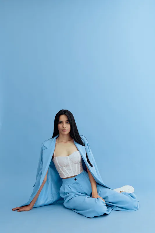 Woman wearing light blue suit and white corset top sitting on the floor against blue background, showcasing modern fashion and serene expression in soft pastel tones.