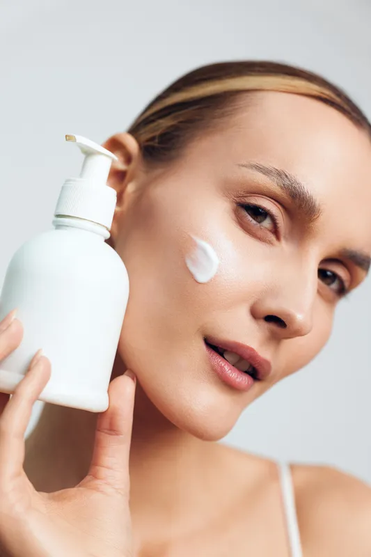 Skincare woman with cream application on cheek holding lotion pump bottle calm beauty routine close portrait on light background natural fresh face care soft studio light hygiene