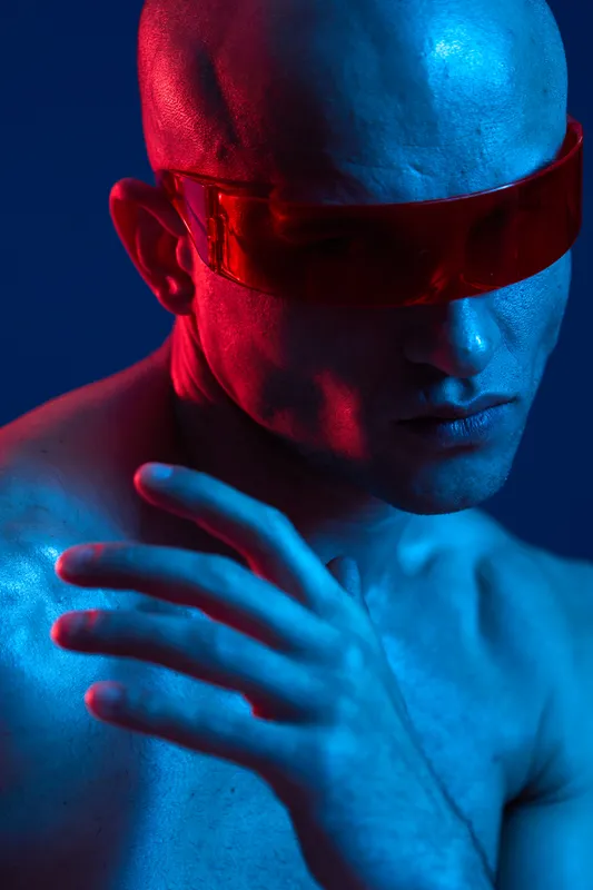 Stylish man with futuristic glasses poses dramatically in vibrant blue and red lighting, embodying a modern and edgy aesthetic. Captivating and unique visual representation.