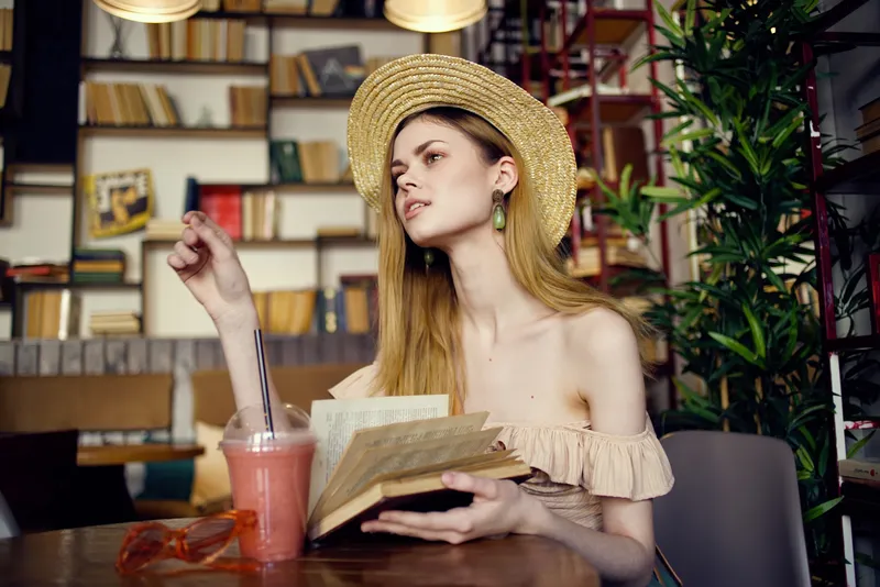 Симпатичная женщина в шляпе с книгой в руках читает, отдыхая в кафе во время отпуска