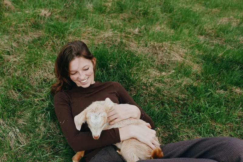Happy relaxed lifestyle shot of a smiling woman resting on grass, cuddling a young goat kid, sharing gentle affection in a sunny outdoor yard, capturing peaceful everyday connection with animals
