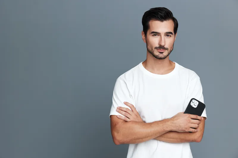 Confident young man in a white t-shirt holding a smartphone, standing with crossed arms against a plain gray background. Casual lifestyle concept.