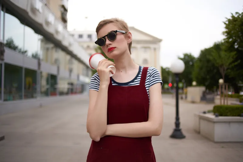 Женщина на улице разговаривает по телефону, в очках, со стаканом напитка в руке, стильная