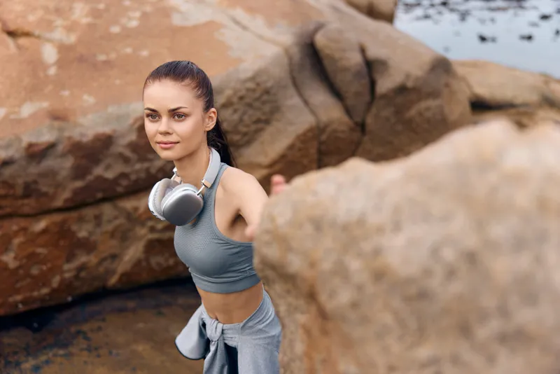 Young woman in sports attire with headphones, enjoying nature by the rocky seaside, exuding confidence and serenity, perfect for lifestyle and fitness themes