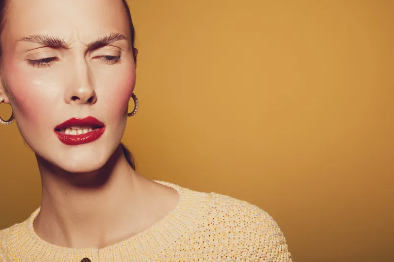 Closeup of a woman with red lipstick and gold earrings making a skeptical face against a warm yellow background. Concept of doubt, emotion, and fashion portrait in studio lighting.