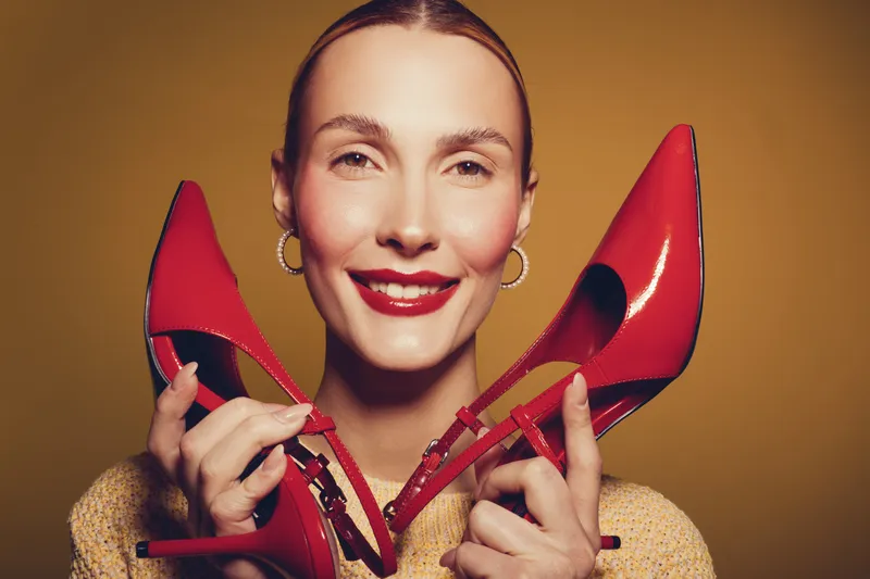 Smiling woman with red lipstick holds red high heel shoes near face on yellow background. Concept of fashion, style, beauty, and confident female model portrait.