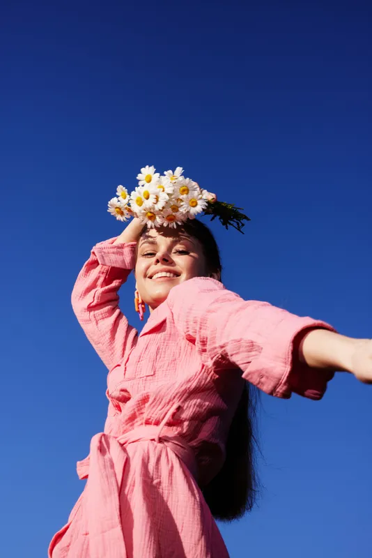 A joyful young woman with long hair wearing a pink dress, playfully posing with daisies against a clear blue sky, embodying a vibrant spring vibe
