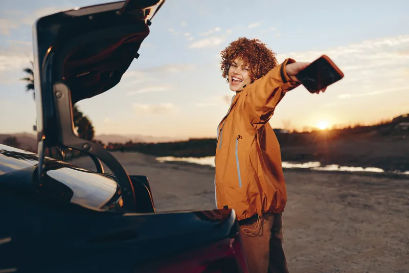 Woman lifestyle outdoors, standing by open car trunk at sunset, holding phone for a warm selfie, smiling with curly hair, casual jacket, calm travel mood, evening light, natural colors