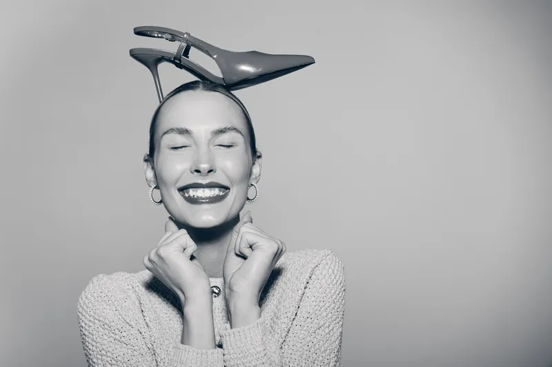 Happy caucasian woman with light skin smiles with closed eyes holding a high heel shoe on her head, wearing cozy sweater and earrings.