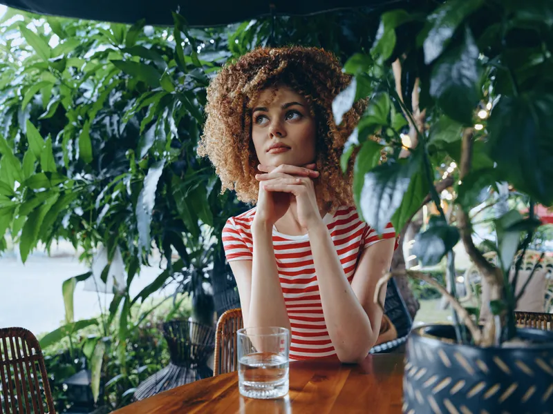 A thoughtful woman with curly hair sits at a wooden table beneath lush green plants, wearing a red striped shirt, resting her chin on hands beside a glass of water.
