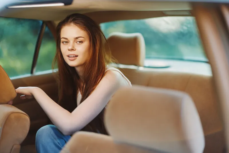 Young woman with long hair sitting in a car, expressing a relaxed and cheerful emotion against a soft-focus green background, capturing a casual moment in nature.