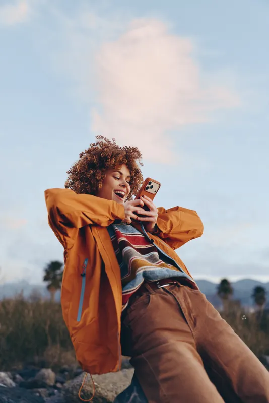 Woman selfie phone outdoors lifestyle smiling curly jacket, a woman in orange windbreaker holds a smartphone for selfie, laughing, hair moves in wind, under blue sky and distant trees, casual