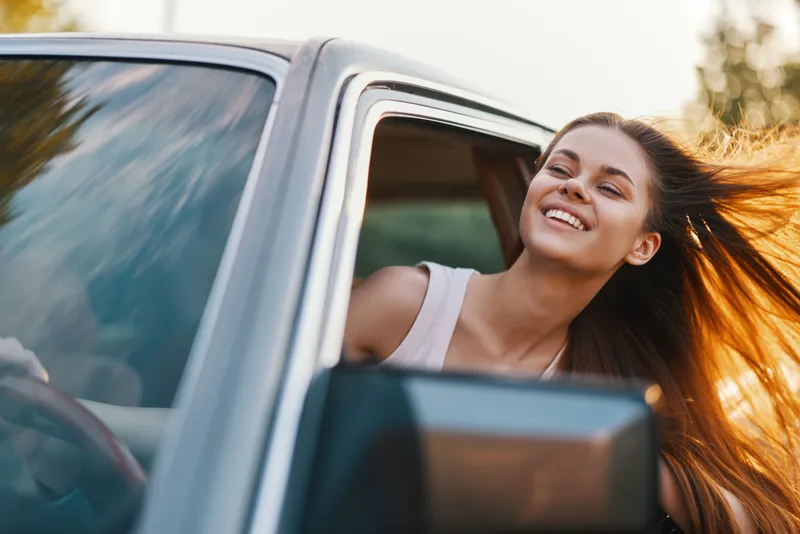 Young woman enjoying freedom and happiness while leaning out of a car window, with long hair flowing in the wind, captured in a vibrant outdoor setting.