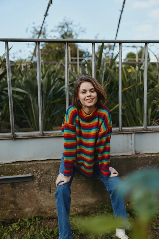 woman smiling sweater colorful casual outdoor sitting jeans on a low wall by metal railing with greenery background, relaxed portrait of young adult in striped knit jumper