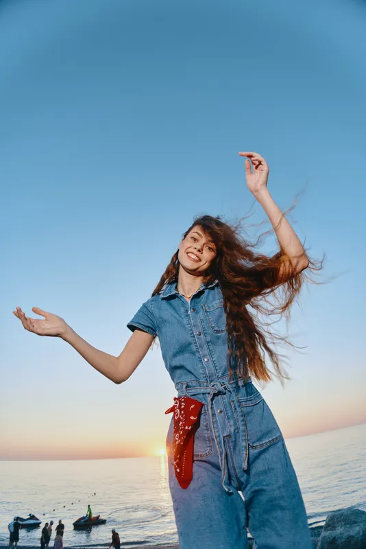Lifestyle woman in modern boho-western denim street style dancing freely by the sea during sunset with film color effect and vibrant natural light.
