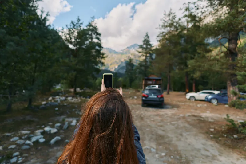 A person with long brown hair raises a smartphone to capture a scenic mountain forest. Seen from behind, the frame conveys exploration, nature, and calm outdoor mood.