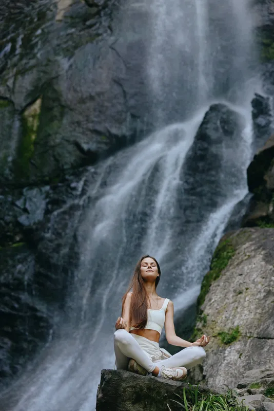 Woman meditating near waterfall in white activewear surrounded by rocks and green moss, showing peaceful nature connection and wellness in outdoor setting.