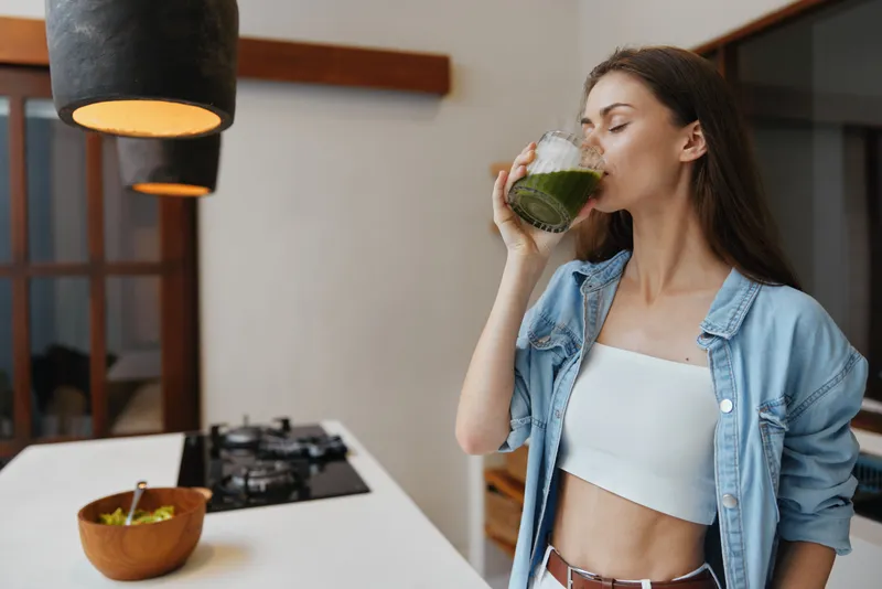 Молодая женщина с длинными волосами в джинсовой куртке пьет зеленый смузи в светлой кухне, пропагандируя здоровый образ жизни и хорошее самочувствие
