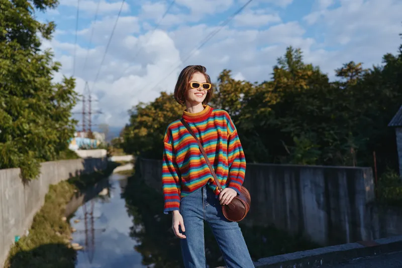 Woman in striped sweater and sunglasses, smiling and leaning on a railing wearing jeans and a crossbody bag beside an urban canal. Young adult casual outfit, travel lifestyle and sunny mood.