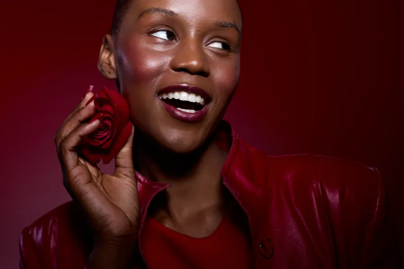 Portrait of a happy African woman with dark skin holding a red rose near her face. She wears a red leather jacket and smiles joyfully against a red background. Concept of beauty and happiness.