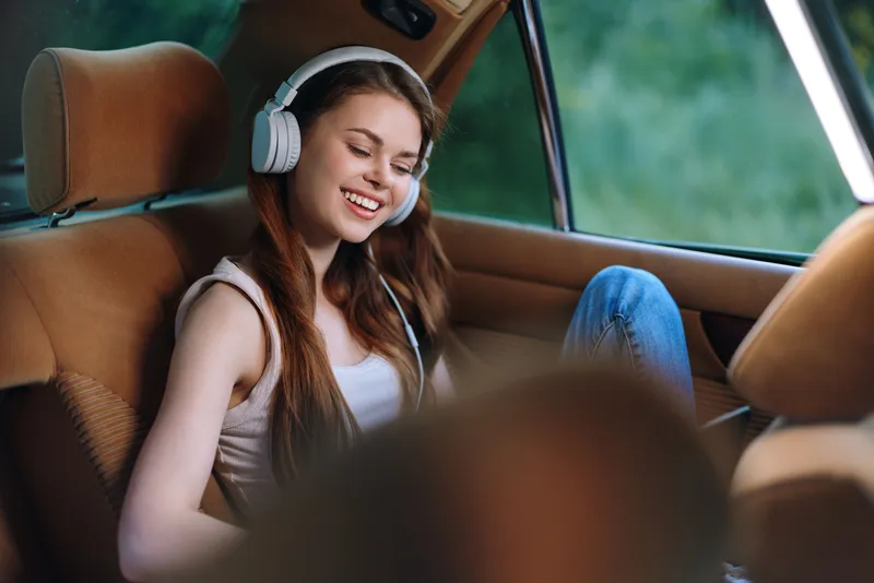 A young woman enjoying music in a car with headphones, smiling joyfully, surrounded by natural light and warm tones. Relaxed atmosphere.