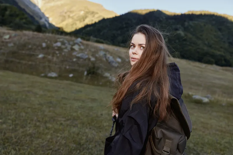 A thoughtful young woman with a backpack stands in an open field with rolling hills and clear blue sky behind her, capturing a serene outdoor moment.