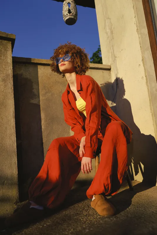Woman in a bright red jumpsuit and yellow top posing on a sunlit terrace, outdoor lifestyle fashion shot focused on confidence, color, and chic style
