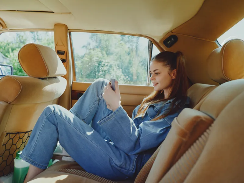 Young woman in casual denim outfit enjoying her smartphone in a cozy car interior, expressing happiness and relaxation during a sunny day trip.