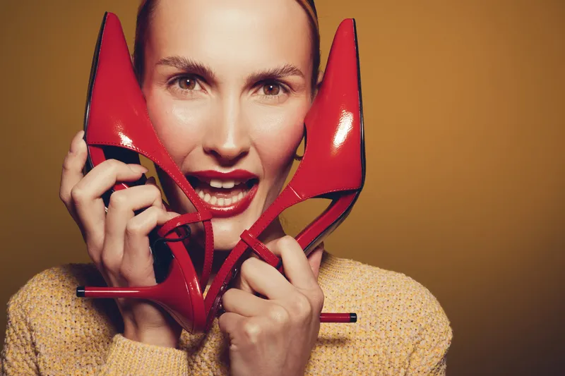 Smiling woman holds red high heel shoes near face, wearing yellow sweater. Studio portrait with warm lighting on brown background. Concept of fashion, style, and footwear.