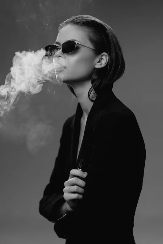 Stylish woman in sunglasses smoking cigarette exhaling smoke in black and white portrait with dark background