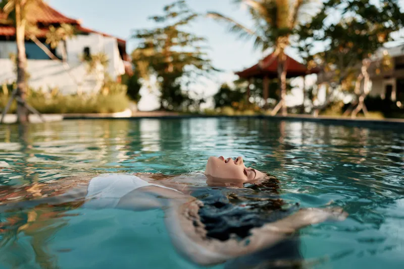 Young woman lifestyle relaxing floating on back in outdoor swimming pool with tropical trees and buildings in the background during sunny day vacation.